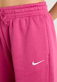 Rozā Nike sporta bikses ar elastīgu jostasvietu un auklu, ar mazu baltu logo sānos. Mīksts audums ar gludu tekstūru.