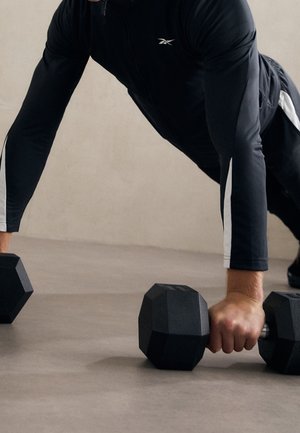 Persoon in zwarte sportkleding die twee zwarte hexagonale dumbbells vasthoudt in een push-up positie op een grijze vloer.