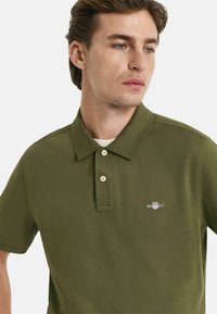 Olivgrön polo med krage och tre knappar. Har en liten broderad logotyp på vänster bröst. Slät yta.