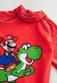 Maglietta rash guard rossa con grafiche di Mario e Yoshi. Tessuto liscio e elastico con collo alto e maniche corte. Accenti di colore a contrasto.