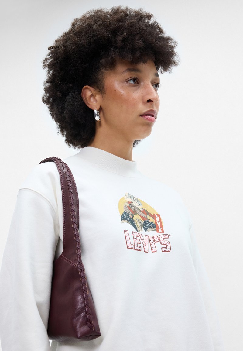 Sudadera blanca con un gráfico colorido de Levi's, combinada con un pequeño bolso de cuero burdeos oscuro con detalles trenzados en la correa.