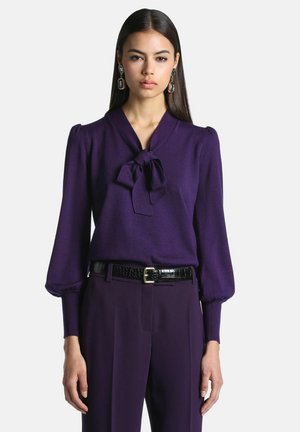 Femme aux longs cheveux foncés portant un chemisier violet avec un nœud au col, un pantalon violet assorti et de grandes boucles d’oreilles rectangulaires.