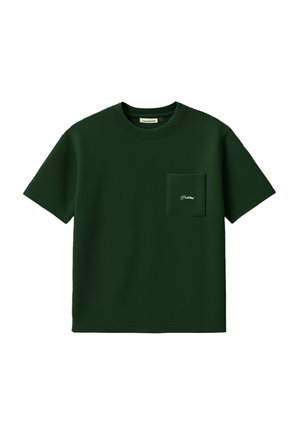 Homme portant une casquette beige, une chemise à manches courtes vert foncé avec poche avant, et un pantalon sombre à fines rayures, debout devant un fond blanc.