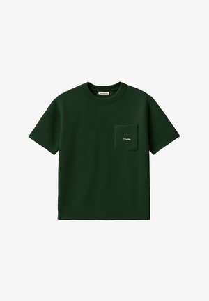 Mand iført beige kasket, mørkegrøn kortærmet skjorte med frontlomme og mørke bukser med striber, står mod hvid baggrund.