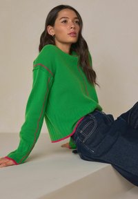 Vihreä neulottu villapaita, jossa on pitsineulottu helma ja vaaleanpunaiset korostukset saumoissa. Yhdistetty tummiin farkkuihin, malli istuu neutraalia taustaa vasten.