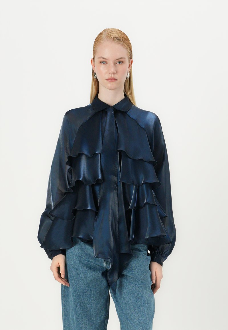 Blouse satin marine avec des volants, col boutonné et manches longues, associée à un jean bleu clair. Texture lisse, design fluide.