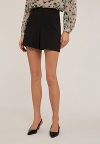 Pantalones cortos negros a medida con dos bolsillos traseros, que presentan un diseño limpio y una textura suave. Se llevan con una blusa floral y tacones de punta.
