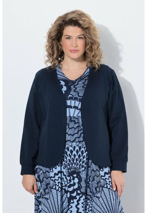 Frau mit lockigen blonden Haaren, die eine marineblaue offene Jacke über ein blau gemustertes Kleid trägt und vor einem schlichten hellen Hintergrund steht.