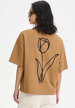 Vrouw draagt een bruin oversized T-shirt met zwarte lijntekening van een bloem op de rug, witte broek en zonnebril, zijwaarts gericht tegen een effen achtergrond.