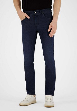 Mann trägt dunkelblaue Slim-Fit-Jeans und weiße Sneaker, linke Hand in der Tasche, steht vor einfachem Hintergrund.