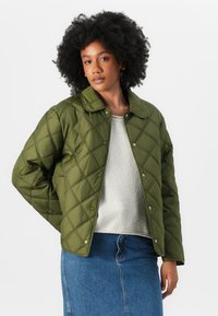 Veste matelassée olive avec col, boutons-pression et motif en losanges, associée à un pull gris clair et une jupe en denim bleu.