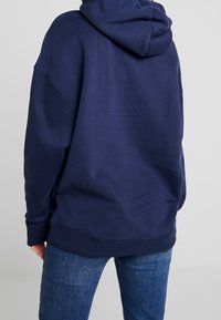 Mörkblå oversized hoodie med dragsko i huvan, ribbade ärmslut och muddar, tillverkad av ett mjukt, texturerat material. Bakvy med minimala detaljer.