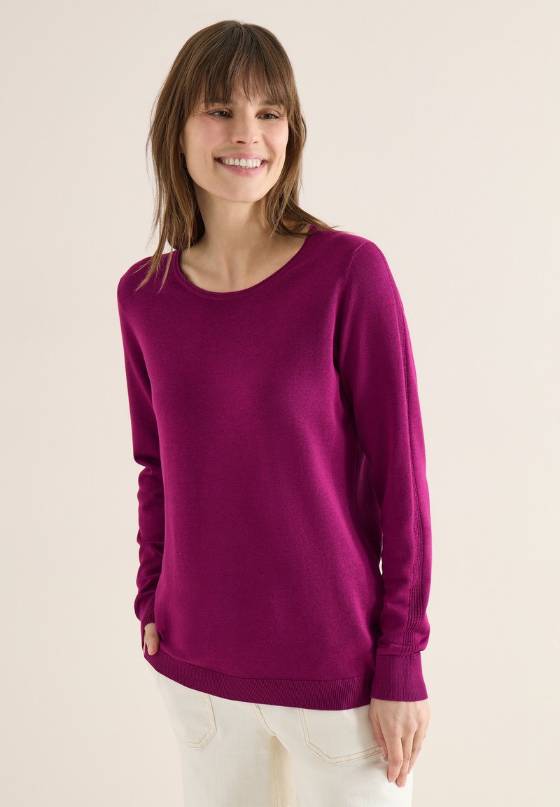 Femme portant un pull à manches longues violet vif et un pantalon beige clair, souriante et regardant légèrement sur le côté devant un fond uni.
