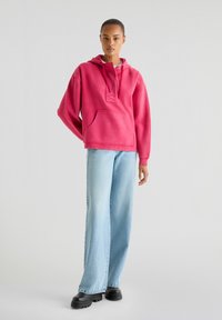 Haut en polaire rose avec une poche avant, un cordon de serrage ajustable et une coupe décontractée, associé à un jean large bleu clair et des bottes à plateforme noires.