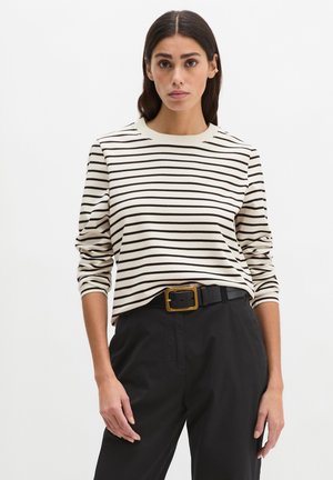 Vrouw draagt een crèmekleurig en zwart gestreept shirt met lange mouwen, ingestopt in high-waisted zwarte broek met een zwarte riem en gouden gesp.