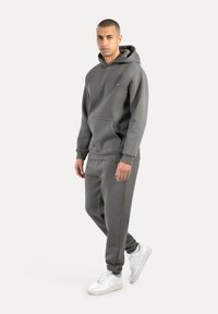 Sudadera gris con capucha y bolsillo frontal, combinada con pantalones jogger a juego. Zapatillas blancas completan el conjunto. Textura suave y diseño casual.