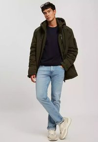 Oliwkowa kurtka puchowa z kapturem, granatowy sweter z okrągłym dekoltem, jasnoniebieskie dżinsy i beżowe sneakersy, charakteryzujące się luźnym krojem i bocznymi kieszeniami.