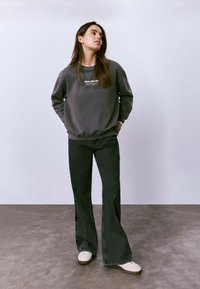 Joven mujer de pie en interiores, vistiendo un sudadera gris oscuro con texto, jeans negros de pierna ancha y zapatillas blancas, con las manos en los bolsillos.