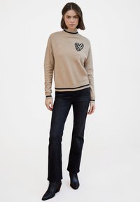 Maglione beige con cuore stampato di leopardo nero sul petto, con collo e polsini a costine, abbinato a jeans a zampa di elefante blu scuro e stivali neri.