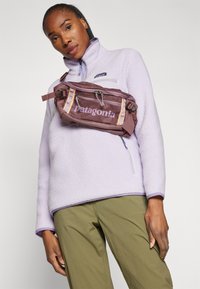 Fleece-pullover i lys lilla, med høy krage og glidelås, kombinert med olivengrønne bukser og en brun Patagonia midjeveske.