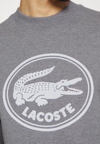Sweat-shirt gris à col rond avec un grand logo blanc Lacoste présentant un crocodile à la bouche ouverte à l'intérieur d'une double bordure ovale.