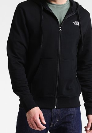 Mand iført en sort hættetrøje med lynlås og The North Face-logo, over en grøn skjorte, kombineret med mørkeblå jeans.