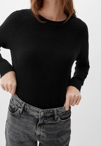 Personne portant un pull noir texturé ajustant la taille d'un jean gris délavé ample sur un fond uni.