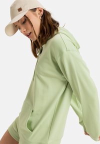 Felpa verde chiaro con tasca frontale e cordini, abbinata a un cappellino beige. Il tessuto sembra morbido, con un design casual e una vestibilità rilassata.