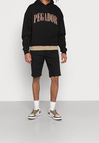 Černá mikina s kapucí s tištěným nápisem "PEGADOR" v růžové, béžová vnitřní vrstva, černé šortky a hnědé/bílé tenisky s texturovanými detaily.