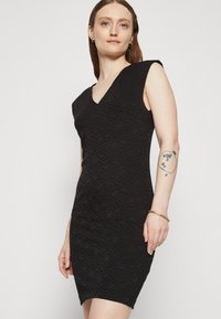 Robe noire à motif avec un décolleté en V, des manches courtes et une silhouette ajustée. Le matériau présente un design géométrique texturé.