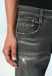 Jeans din denim gri, cu o textură uzată, dungi subțiri și o mică ruptură. Buzunarul din spate are un design distinct în formă de V.