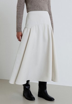 Donna che indossa una lunga gonna bianca a ruota, un maglione beige in maglia e stivali neri in pelle al ginocchio, in piedi su un pavimento chiaro contro una parete bianca.