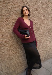 Bordowy sweter z dekoltem w serek i złotymi guzikami, w połączeniu z przezroczystą czarną spódnicą, trzymając czarną skórzaną torbę, na tle teksturowanej ściany.