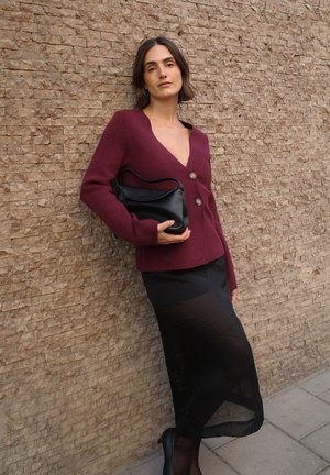 Bordový pletený sveter s výstrihom do V a zlatými gombíkmi, v kombinácii s priesvitnou čiernou sukňou, s čiernou koženou taškou, pred textúrovanou stenou.