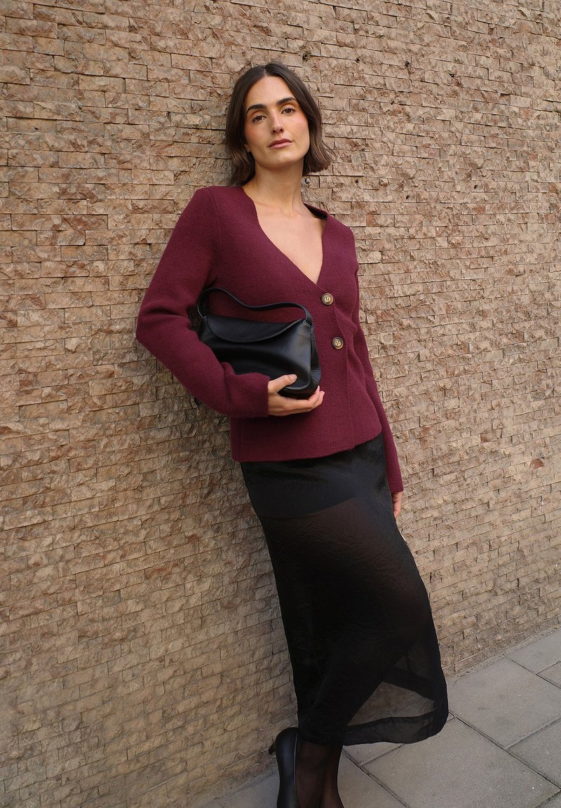 Bordowy sweter z dekoltem w serek i złotymi guzikami, w połączeniu z przezroczystą czarną spódnicą, trzymając czarną skórzaną torbę, na tle teksturowanej ściany.