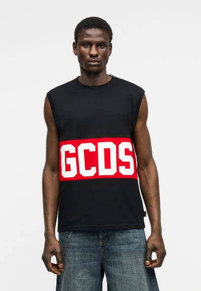 Νεαρός άνδρας με μαύρο αμάνικο τοπ με κόκκινη λωρίδα και λευκά γράμματα "GCDS", συνδυασμένο με φαρδιά μπλε τζιν.