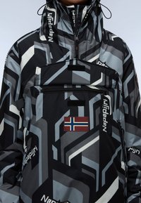 Personne portant une veste Napapijri à motifs noirs, gris et blancs, avec une poche zippée à l'avant et un écusson du drapeau norvégien sur la poitrine.