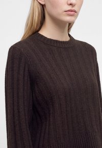 Femme aux longs cheveux blonds portant un pull en maille côtelée marron foncé avec un col rond sur un fond clair uni.
