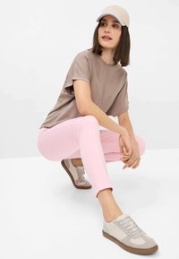 Camicia a maniche corte color tan, jeans aderenti rosa chiaro, sneakers grigie e rosa con accenti crema, e un cappellino beige con rifiniture blu.