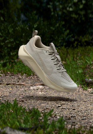 Μπεζ παπούτσι για τρέξιμο σε μονοπάτι Nike στον αέρα πάνω από χαλικώδη μονοπάτι με φόντο πράσινα φύλλα, που αναδεικνύει την υφή της σόλας και το διαπνέον άνω μέρος από δίχτυ.