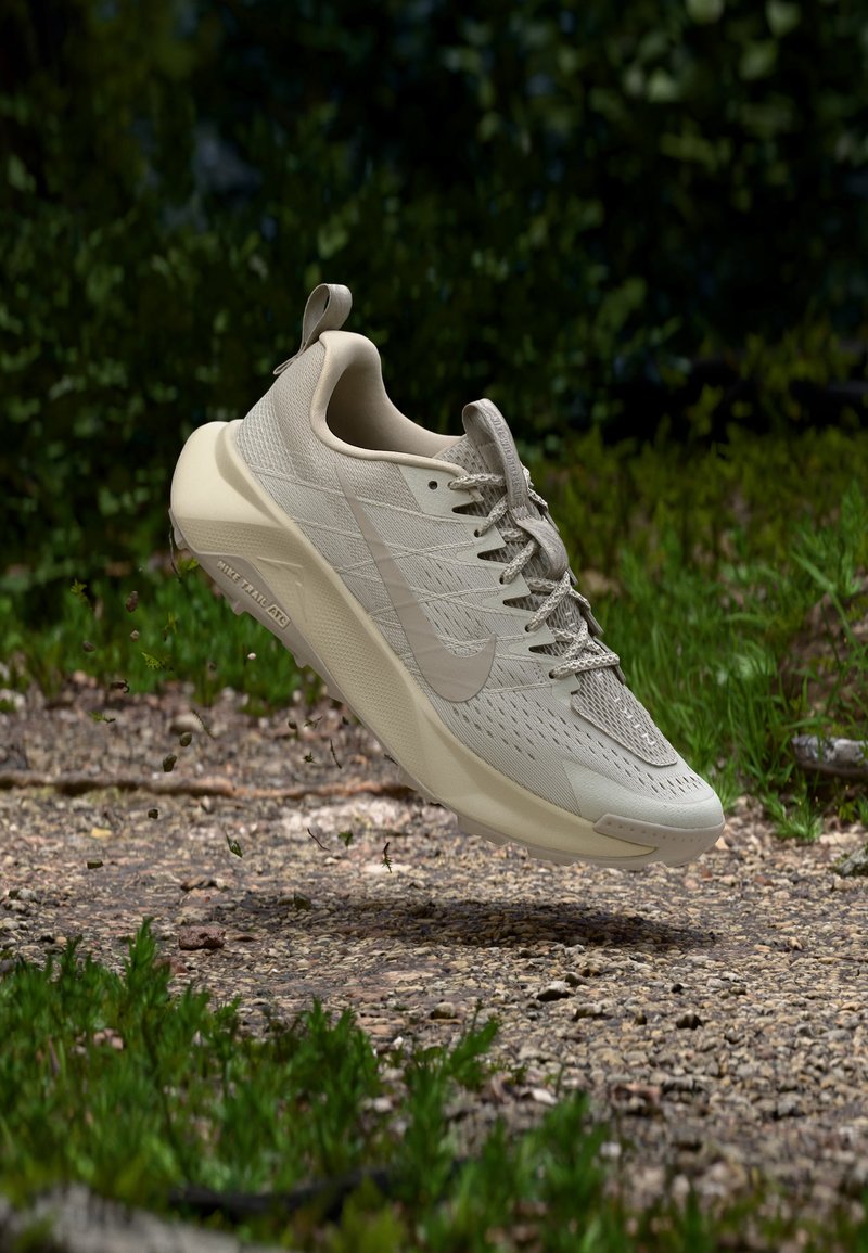Beige Nike terrengløpesko midt i lufta over grussti med grønt løvverk i bakgrunnen, med fokus på teksturert såle og pustende mesh-overdel.