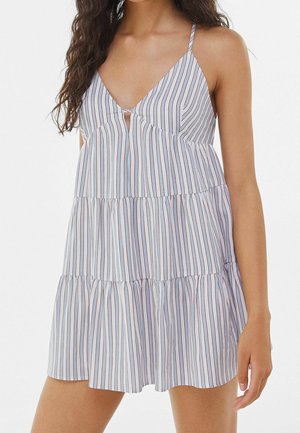 Femme portant une mini robe à volants sans manches avec des bretelles fines et des rayures verticales bleues, blanches et rouges, montrant le haut du corps et le bras droit.