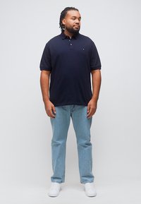 Άνδρας με πλεγμένα μαλλιά που φοράει μπλε πουκάμισο πόλο, ανοιχτό μπλε τζιν και λευκά αθλητικά παπούτσια, όρθιος μπροστά σε ένα απλό ανοιχτόχρωμο φόντο.