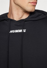 Felpa nera con cappuccio in tessuto morbido, dotata di tasca frontale, cappuccio con cordini e logo bianco "HUMMEL" sul petto.