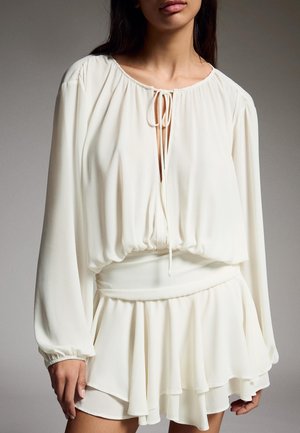 Woman wearing a long-sleeve, cream-colored blouse with a tie at the neckline and a matching layered skirt, standing against a plain background.