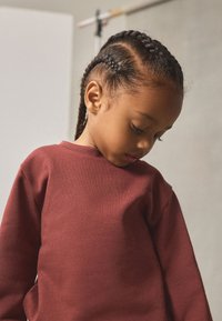 Sweat-shirt bordeaux avec un col rond et des poignets côtelés, porté par un enfant aux cheveux tressés, regardant la texture du tissu.