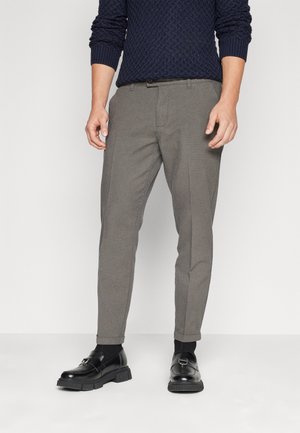 Mann trägt graue Slim-Fit-Hose, schwarze Socken, schwarze Leder-Loafer und einen dunkelblauen, strukturierten Pullover, steht vor weißem Hintergrund.