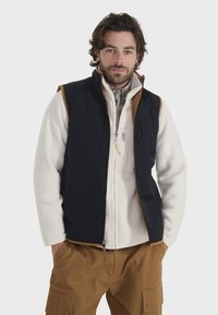 Chaleco negro sin mangas sobre una chaqueta de forro polar blanca, con cierre frontal, acentos marrones y pantalones cargo marrones. Textura suave, diseño funcional.