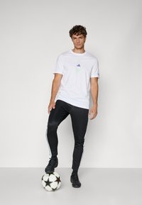 T-shirt blanche avec un logo graphique et un accent vert, assortie à un pantalon de jogging noir avec des rayures blanches sur les côtés. Un ballon de football noir est posé au sol.