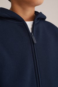 Svetlo modra zip-up kapuca izdelana iz mehke tkanine z gladko teksturo. Ima srebrn zadrgo in kapuco. Brez vidnih vzorcev ali okrasnih elementov.